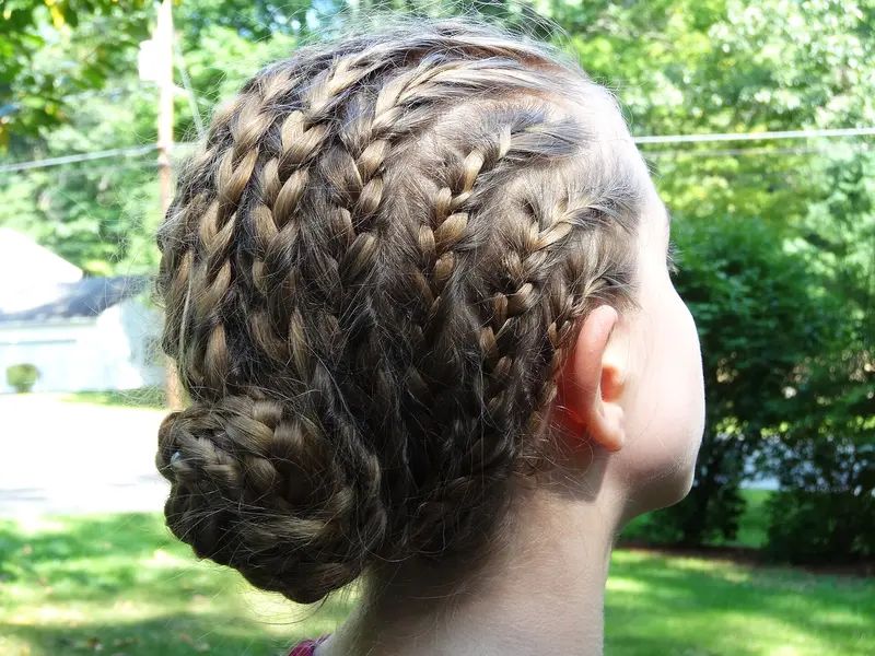 Back of a head with light brown hair in several neat braids running from the hairline and twisted into a low braided bun at the nape, against a sunny green garden