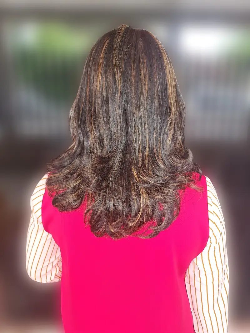 Seen from behind, a woman's shoulder-length dark brown hair with caramel highlights falls in soft layered waves over a bright pink top