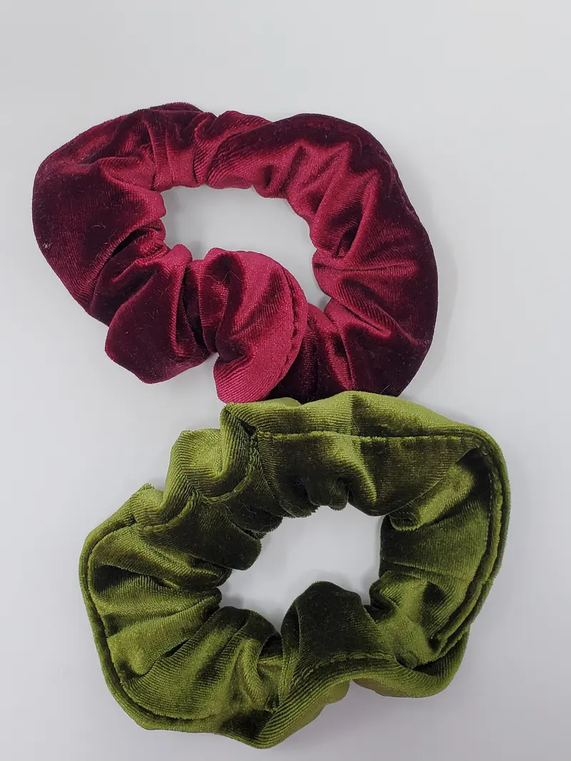 Two soft velvet scrunchies, one deep burgundy and one olive green, lying on a pale grey background
