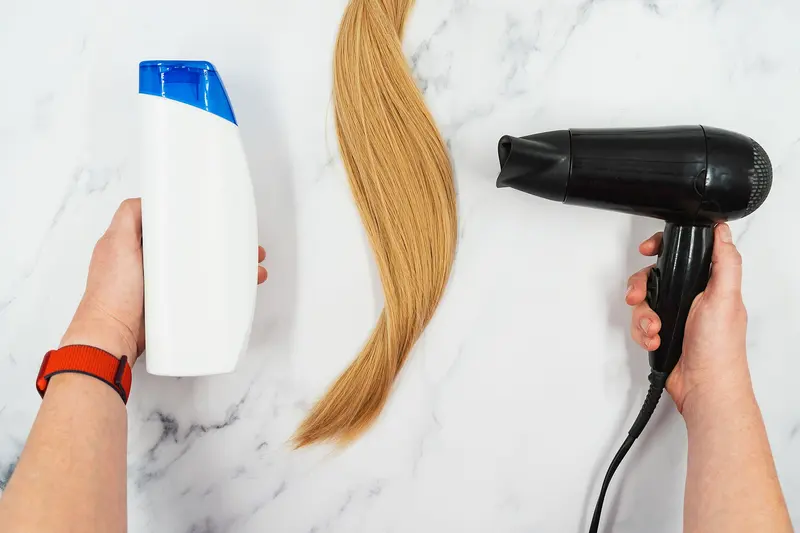 Flat lay on white marble: one hand holds a black hairdryer, the other a plain white shampoo bottle, with a long lock of honey-blonde hair between them