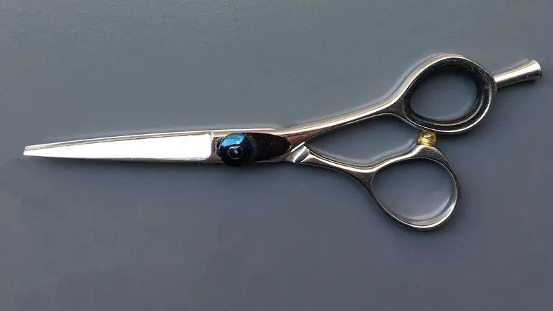 A pair of polished stainless-steel hairdressing shears with offset finger rings and a finger rest, lying flat on a matt slate-grey surface