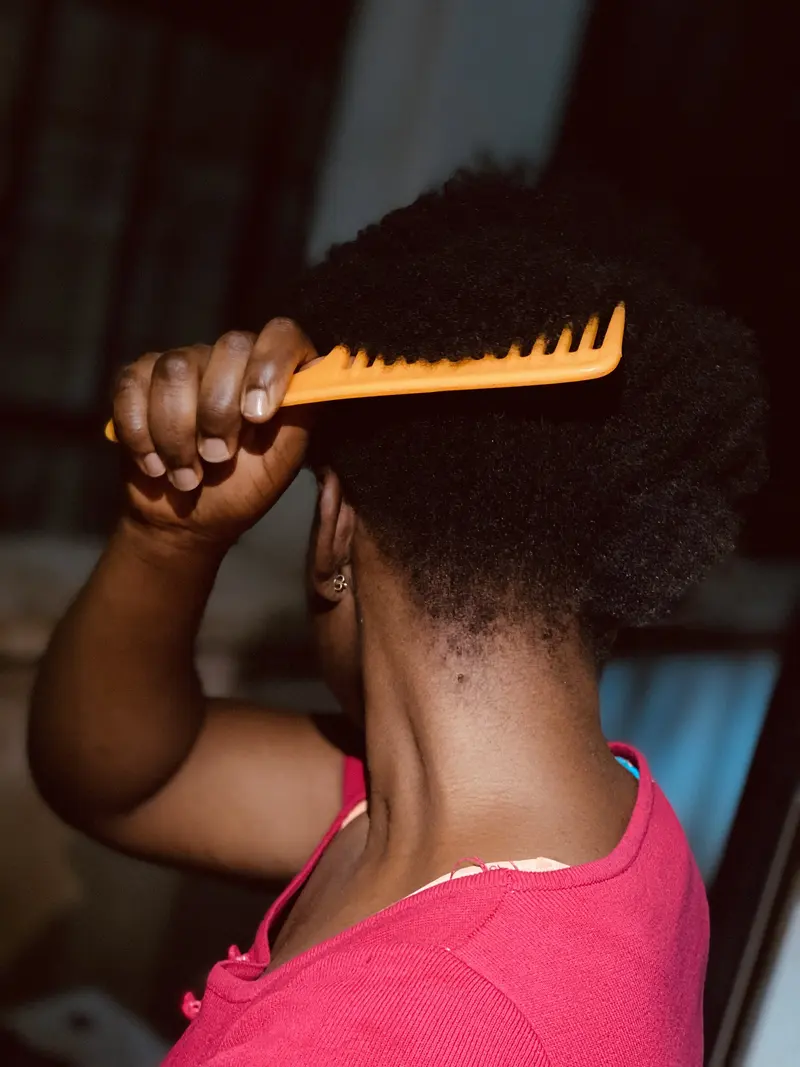 Seen from behind, a woman in a pink top lifts an orange wide-tooth comb through her short, dense afro-textured hair against a dim background